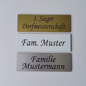 Könnte beinhalten: Drei rechteckige Plaketten in Gold, Weiß und Silber mit schwarzem Text. Die obere Plakette lautet "1. Sieger Dorfmeisterschaft". Die mittlere Plakette lautet "Fam. Muster". Die untere Plakette lautet "Familie Mustermann".