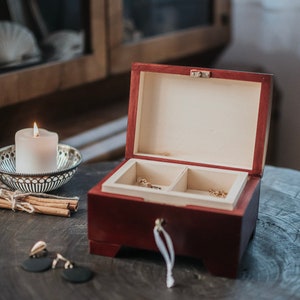 May include: A rectangular wooden jewelry box with a dark reddish-brown exterior and a light cream-colored interior featuring two compartments. The box has a small metal latch and a white ribbon pull. Nearby are cinnamon sticks and a white candle.