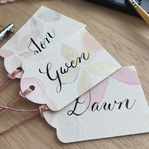 May include: Three white gift tags with pink and gold watercolor designs. The tags are tied with pink string and have the names "Jon", "Gwen", and "Dawn" written in black calligraphy.
