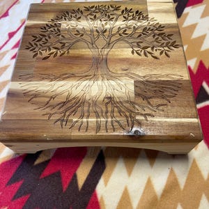 May include: A square wooden riser with a natural grain finish, featuring a detailed laser-engraved design of a tree with expansive branches and visible roots centered on the top surface. The riser stands on four short, sturdy feet.