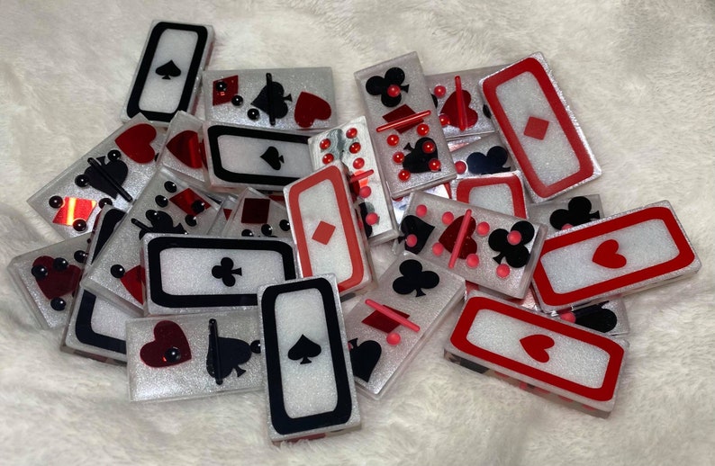 May include: A pile of white and red dominoes with black and red suit symbols. The dominoes are decorated with glitter and have a white background with red and black borders.