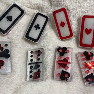 May include: A set of ten dominoes with a white background and black and red accents. The dominoes feature playing card symbols, including hearts, spades, clubs, and diamonds. The dominoes are made of a clear material with a glittery finish.