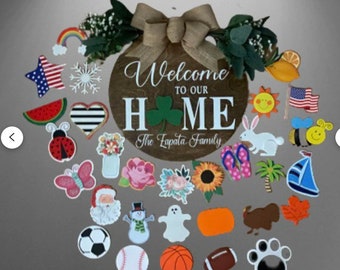 May include: A wooden round sign with the text "Welcome to our Home The Zapata Family" surrounded by a variety of colorful wooden cutouts, including a ladybug, a butterfly, a snowman, a ghost, a sunflower, a watermelon, a sailboat, a palm tree, a heart, a star, a snowflake, a sun, a bee, a rabbit, a pair of flip flops, a pumpkin, a turkey, a leaf, a soccer ball, a baseball, a basketball, a football, and a paw print.