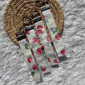May include: Five fabric keyrings with a white background, patterned with red strawberries and blue flowers. Each keyring has a silver metal clasp and ring. The keyrings are arranged on a woven, circular placemat.