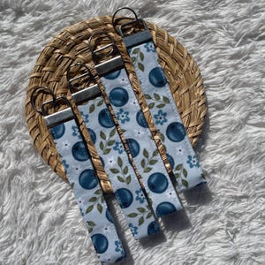 May include: Four fabric keychains with a light blue backdrop, patterned with dark blue blueberries, white flowers, and green leaves. Each keychain has a silver metal ring and a matching fabric loop.