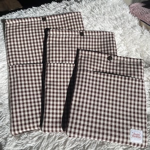 May include: A set of three brown and white gingham fabric pouches of varying sizes. Each pouch has a pocket and a snap closure. The smallest pouch has a label that reads "June Flower."