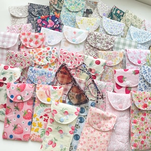 Puede incluir: Una colección de estuches de tela coloridos para gafas con varios patrones florales y de frutas, incluyendo fresas y cerezas. Los estuches se cierran con un botón y presentan una variedad de colores, incluyendo rosa, azul y verde. Dos pares de gafas son visibles.