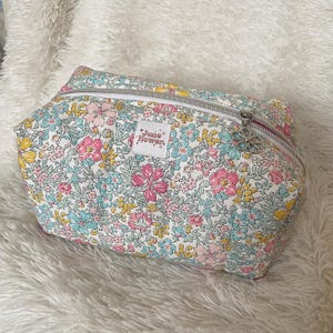 Peut inclure: Une trousse de maquillage à imprimé floral avec un fond blanc, ornée de fleurs roses, jaunes et bleues. Le sac a une fermeture éclair argentée et une tirette en forme de fleur. L'étiquette indique "June Flower."