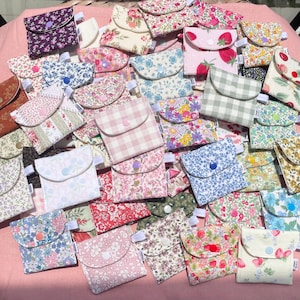 May include: A selection of small fabric pouches, featuring floral, gingham, and fruit designs. Each pouch has a curved flap fastened with a button. The pouches are in pink, blue, green, and white, creating a colourful display.