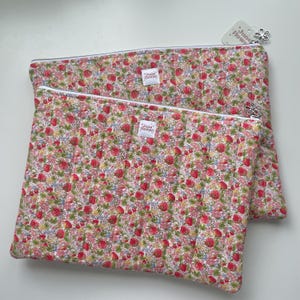 May include: Two rectangular quilted pouches featuring a dense floral pattern of red, pink, and green on a white background. Each pouch has a white zip with a silver flower-shaped pull and a small white label reading June Flower in the centre.