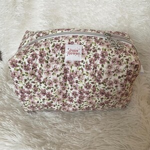 May include: A floral print cosmetic bag with a silver zip. The bag features a pattern of small purple flowers and green leaves on a white background. The label reads "June Flower."