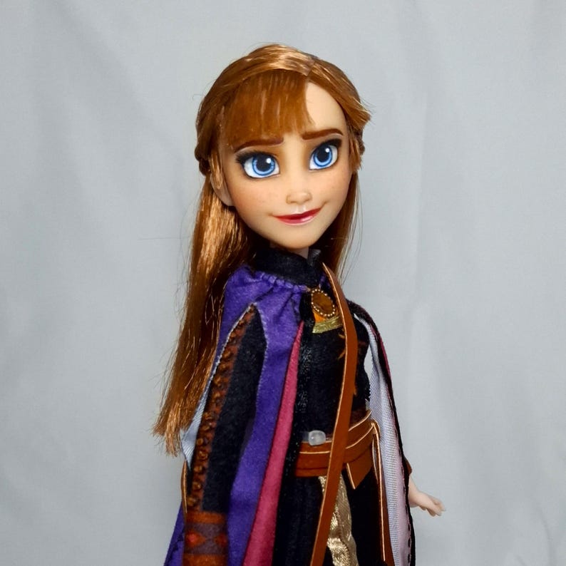 May include: A doll of a character with auburn hair, blue eyes, and a smiling expression. The doll wears a black dress with a purple and pink cape, and a brown belt. The doll is a collectible item.