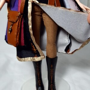 May include: A doll dressed in a detailed costume, featuring a brown leather satchel, belt, and boots. The outfit includes a black coat with gold trim and a colorful inner lining. The doll's legs are clad in brown leggings.