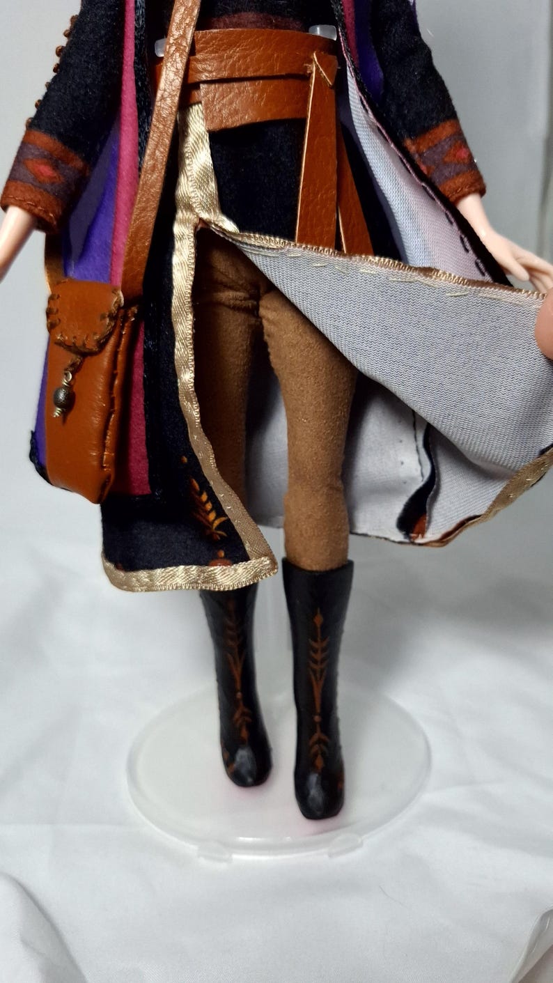 May include: A doll dressed in a detailed costume, featuring a brown leather satchel, belt, and boots. The outfit includes a black coat with gold trim and a colorful inner lining. The doll's legs are clad in brown leggings.