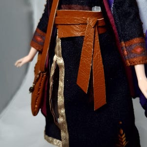 May include: A doll wearing a black dress with gold trim and a brown leather belt and shoulder bag. The dress has orange and yellow leaf-like designs. A purple cape with red and blue accents is draped over the shoulders.