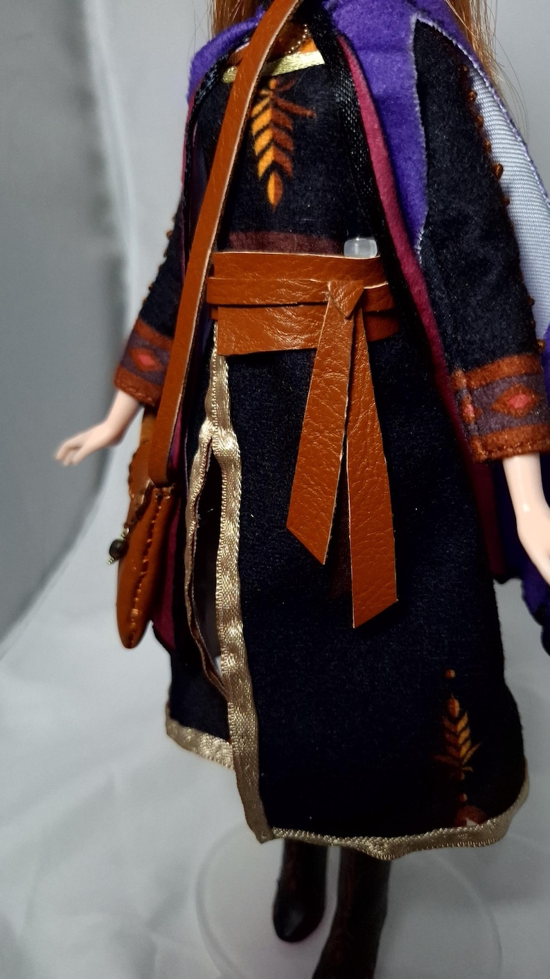 May include: A doll wearing a black dress with gold trim and a brown leather belt and shoulder bag. The dress has orange and yellow leaf-like designs. A purple cape with red and blue accents is draped over the shoulders.