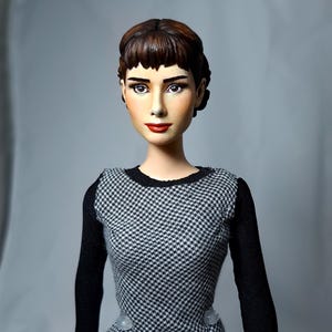 May include: A doll with a detailed face and brown hair styled in an updo with bangs. The doll wears a black and white houndstooth dress with long black sleeves. The doll's lips are painted red, and it has a neutral background.