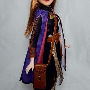 May include: A doll of a character with auburn hair and blue eyes, wearing a black dress with a purple cape and brown accessories. The doll has a small brown purse and black boots. The doll is standing on a clear plastic stand.
