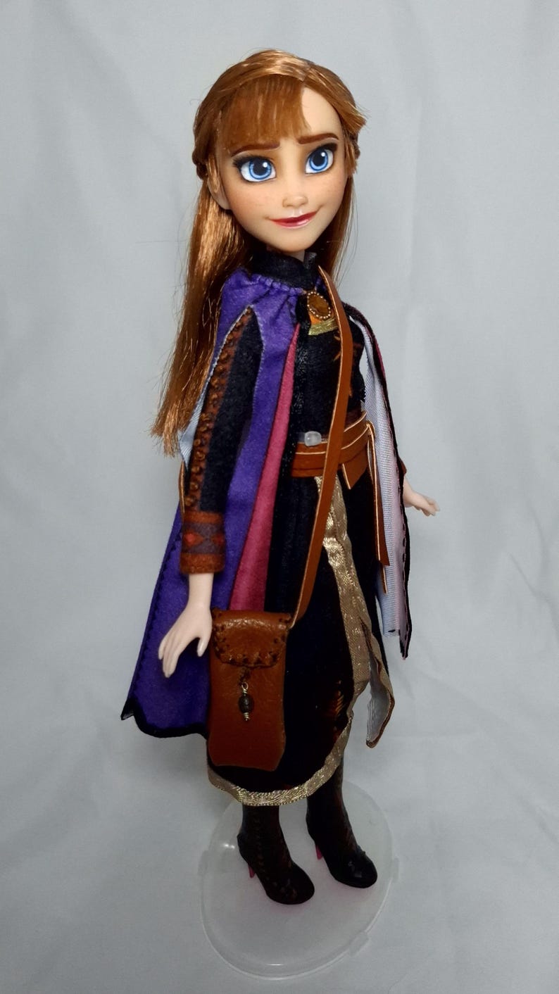 May include: A doll of a character with auburn hair and blue eyes, wearing a black dress with a purple cape and brown accessories. The doll has a small brown purse and black boots. The doll is standing on a clear plastic stand.