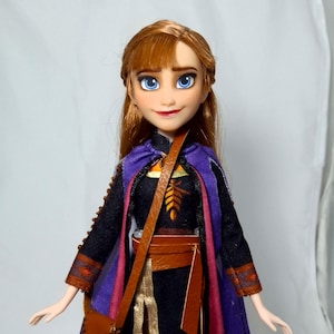 May include: A doll of a character with auburn hair and blue eyes, wearing a black dress with a purple cape and brown satchel. The doll has a smiling expression and is a collectible item.