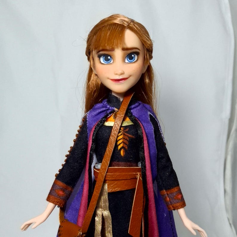 May include: A doll of a character with auburn hair and blue eyes, wearing a black dress with a purple cape and brown satchel. The doll has a smiling expression and is a collectible item.