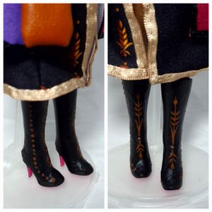 May include: Close-up of a doll's legs and lower torso. The doll wears black boots with gold accents and pink heels. The doll's outfit includes a black skirt with gold trim and a purple, orange, and black bodice.