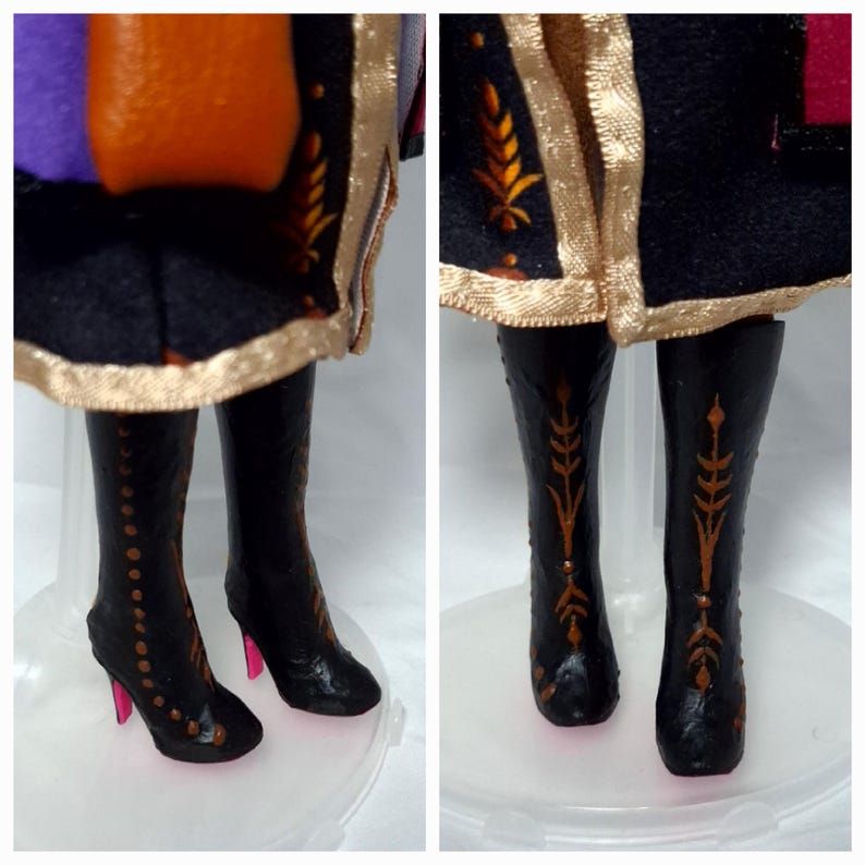 May include: Close-up of a doll's legs and lower torso. The doll wears black boots with gold accents and pink heels. The doll's outfit includes a black skirt with gold trim and a purple, orange, and black bodice.