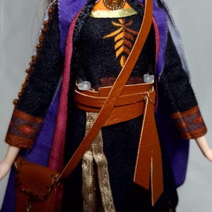 May include: A doll wearing a black dress with orange and brown accents, a purple cape, and a brown leather satchel. The doll has red hair and is wearing a gold-colored necklace. The doll's outfit has gold trim.