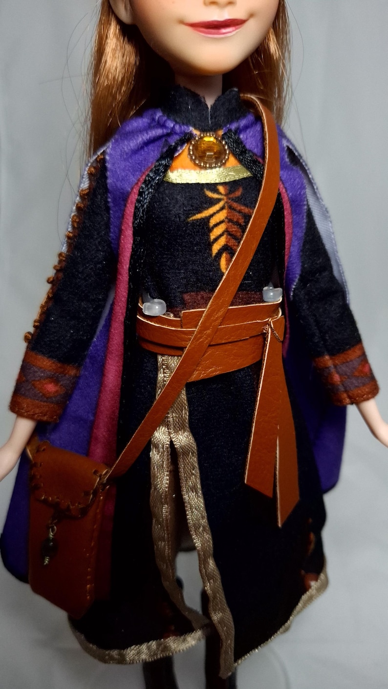 May include: A doll wearing a black dress with orange and brown accents, a purple cape, and a brown leather satchel. The doll has red hair and is wearing a gold-colored necklace. The doll's outfit has gold trim.