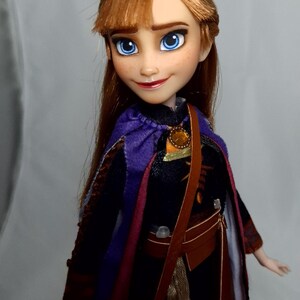 May include: A doll of a character with auburn hair, blue eyes, and a purple cape. The doll wears a dark dress with a brown satchel and a gold-colored pendant. The doll's face has freckles and a slight smile.