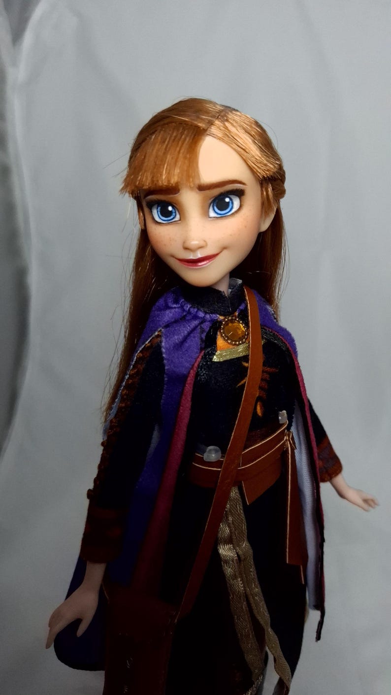 May include: A doll of a character with auburn hair, blue eyes, and a purple cape. The doll wears a dark dress with a brown satchel and a gold-colored pendant. The doll's face has freckles and a slight smile.