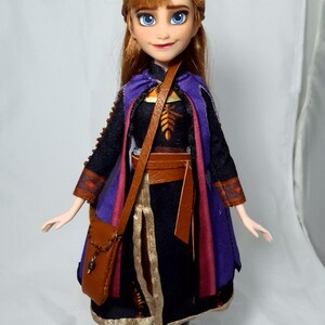 May include: A doll of a character with auburn hair and blue eyes, wearing a black dress with a purple and red cape, brown belt, and a brown satchel. The doll is standing on a clear plastic stand.