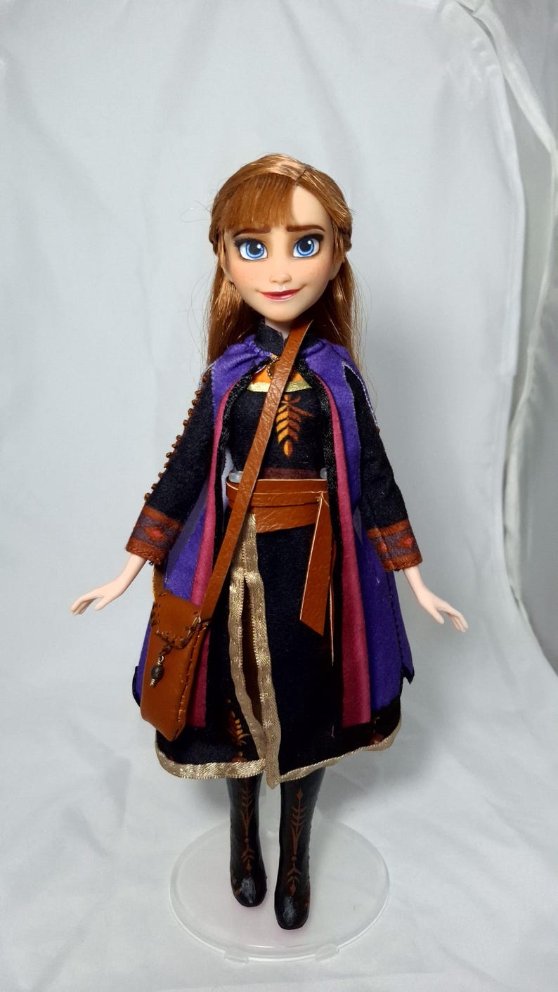 May include: A doll of a character with auburn hair and blue eyes, wearing a black dress with a purple and red cape, brown belt, and a brown satchel. The doll is standing on a clear plastic stand.
