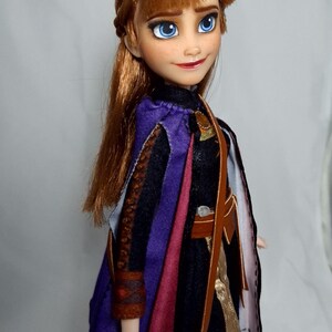 May include: A doll of a character with auburn hair, blue eyes, and a red lip color. The doll wears a black and purple outfit with a brown leather bag. The doll's outfit includes a purple cape and a brown leather bag.