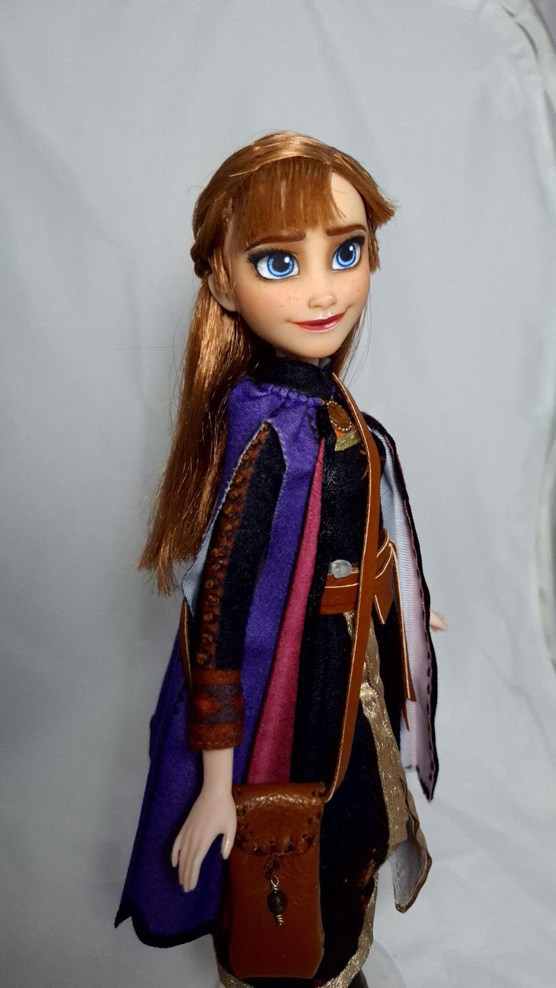 May include: A doll of a character with auburn hair, blue eyes, and a red lip color. The doll wears a black and purple outfit with a brown leather bag. The doll's outfit includes a purple cape and a brown leather bag.