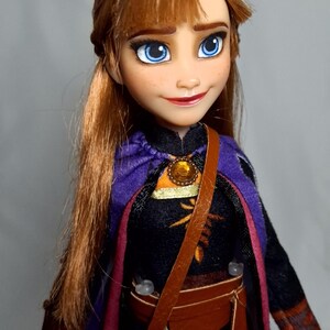 May include: A doll of a character with auburn hair and blue eyes, wearing a black and purple outfit with a brown satchel. The doll has a detailed face with freckles and a small smile. The outfit includes a purple cape and a brown belt.