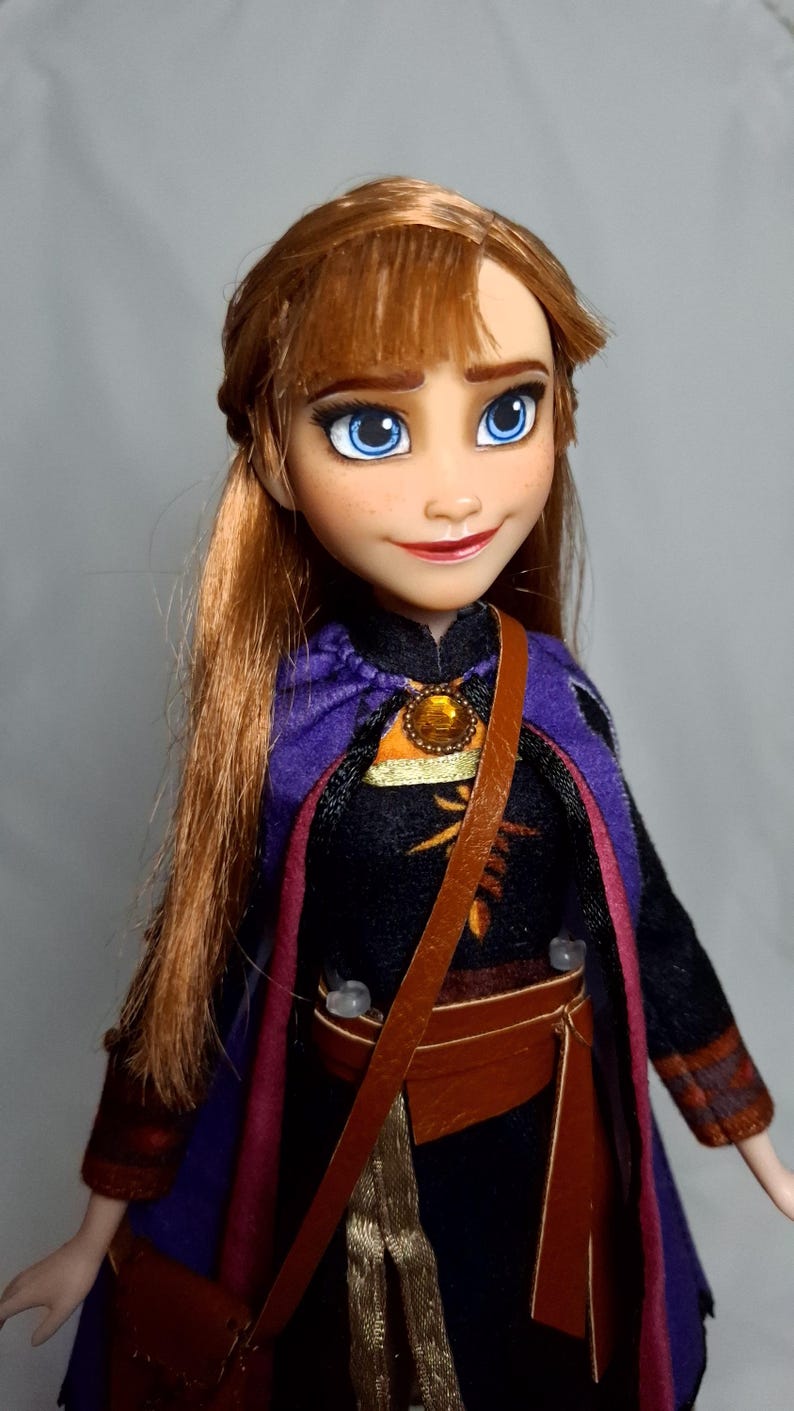 May include: A doll of a character with auburn hair and blue eyes, wearing a black and purple outfit with a brown satchel. The doll has a detailed face with freckles and a small smile. The outfit includes a purple cape and a brown belt.