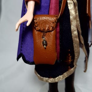 May include: A doll wearing a purple and black outfit with a brown leather shoulder bag. The bag has a decorative bead and a strap. The doll's outfit includes a cape and a gold trim.