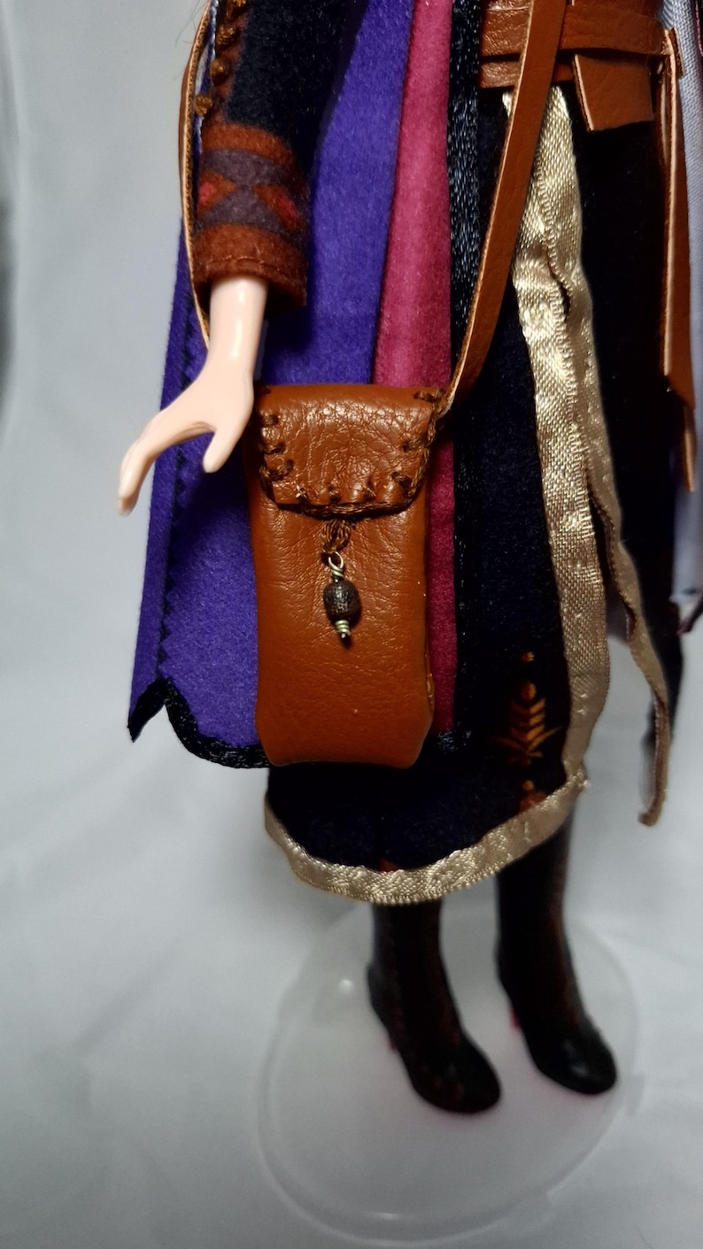 May include: A doll wearing a purple and black outfit with a brown leather shoulder bag. The bag has a decorative bead and a strap. The doll's outfit includes a cape and a gold trim.