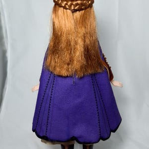 May include: A doll with long, auburn hair styled with a braided crown and flowing down the back. The doll wears a purple cape with black trim and black boots. The doll is standing on a clear plastic stand.