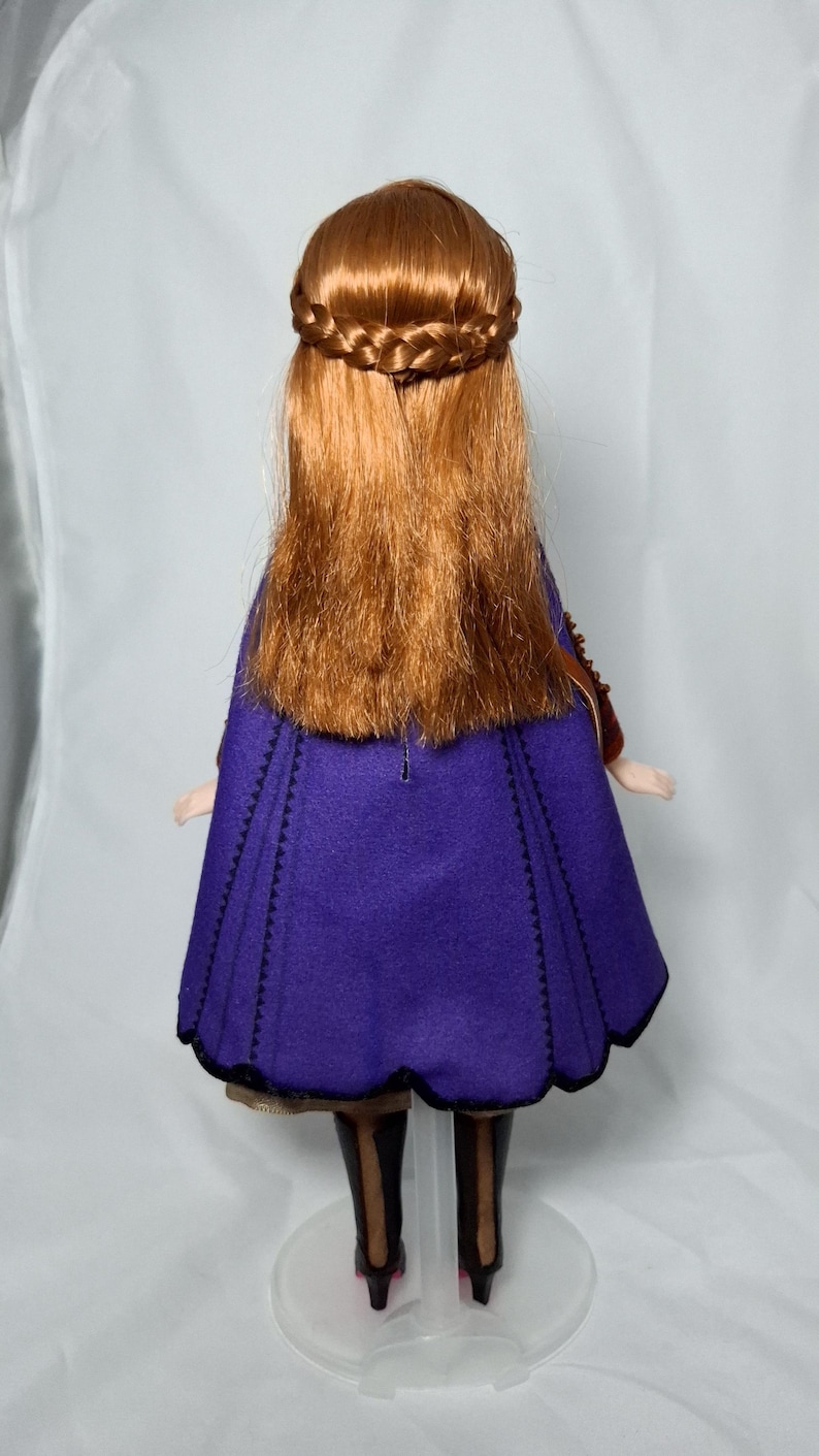 May include: A doll with long, auburn hair styled with a braided crown and flowing down the back. The doll wears a purple cape with black trim and black boots. The doll is standing on a clear plastic stand.