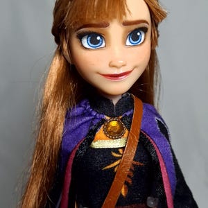 May include: A doll of a character with auburn hair, blue eyes, and a smiling expression. The doll wears a black dress with a purple cape and a brown leather strap across the chest. A gold pendant is visible.