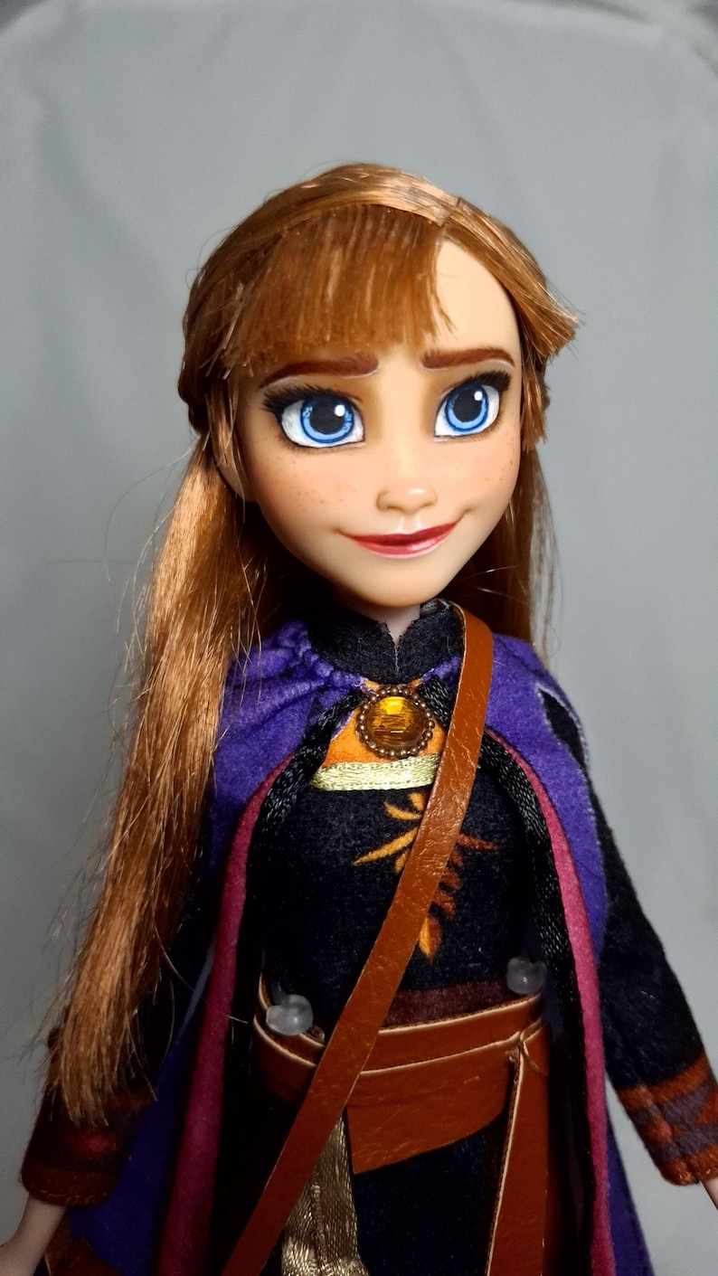 May include: A doll of a character with auburn hair, blue eyes, and a smiling expression. The doll wears a black dress with a purple cape and a brown leather strap across the chest. A gold pendant is visible.