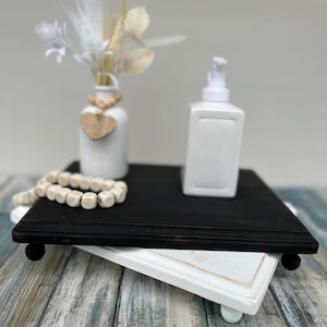 May include: A black and white tiered tray with a white vase filled with dried flowers, a white soap dispenser, and a wooden bead garland. The tray is sitting on a wooden surface.