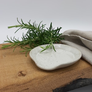 Könnte beinhalten: Ein kleiner, gesprenkelter weißer Keramik-Löffelhalter mit organischer Form, auf einem Holzbrett. Frische Rosmarinzweige und ein gefaltetes beiges Leinentuch verleihen eine natürliche Note. Ideal für die Küche.