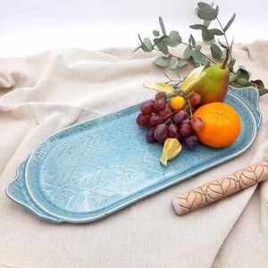 Könnte beinhalten: Eine hellblaue Keramik-Servierplatte mit einem Blatt-Muster. Die Platte hat zwei Griffe und ist mit Obst gefüllt, darunter Trauben, eine Birne und eine Orange.