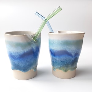 Könnte beinhalten: Zwei Keramikbecher mit blau-weißem Farbverlauf, jeweils mit einem Glasstrohhalm. Die Becher haben eine einzigartige Form und einen tropfenden Glanzeffekt am Boden. Ein Strohhalm ist grün, der andere blau.