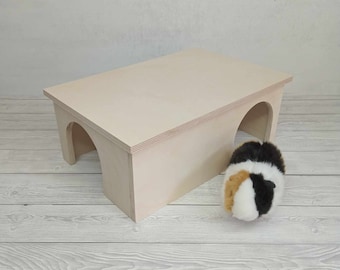 Op de afbeelding: Een licht houten schuilplaats met twee tunnels voor kleine huisdieren, zoals hamsters of cavia's. De schuilplaats heeft een plat dak en twee gebogen ingangen.