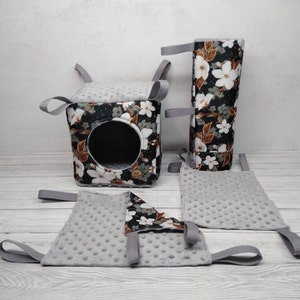 Puede incluir: Un conjunto de cuatro accesorios colgantes para hámster con estampado floral. El conjunto incluye una casa en forma de cubo, un tubo y dos plataformas colgantes. La casa y el tubo tienen un fondo negro con flores blancas y detalles marrones. Las plataformas son grises con una superficie texturizada.