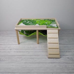May include: A light wood pet bed with a green and white leaf-patterned cushion. The bed has a built-in ladder and a hammock underneath. The bed is designed for small pets.